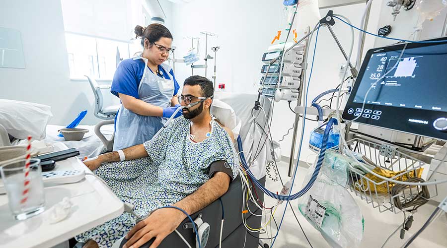 Healthcare worker beside patient hooked to medical devices in hospital room.