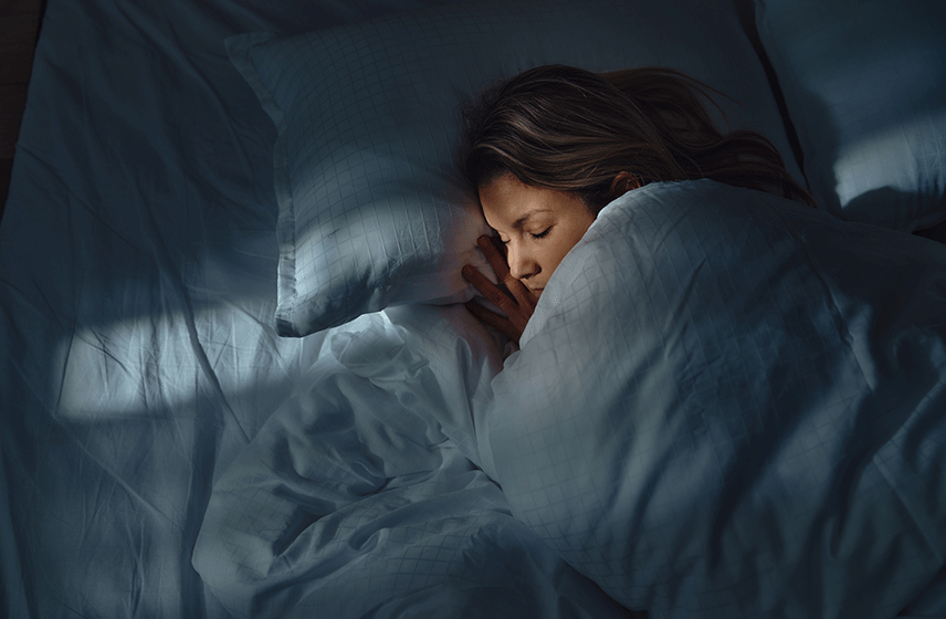 Person sleeping in bed with their face partially obscured by a pillow and bed sheets.